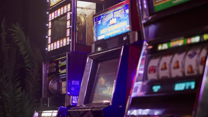 The video shows a closeup of a row of slot machines inside a dimly lit casino or arcadethe machines are brightly lit featuring colorful game screens but the overall atmosphere of the room is dark creating a strong contrast between the machine