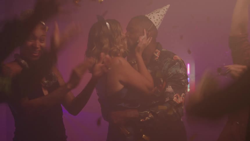 Some people are happy and wearing festive hats and then golden papers fall on two people who are kissing