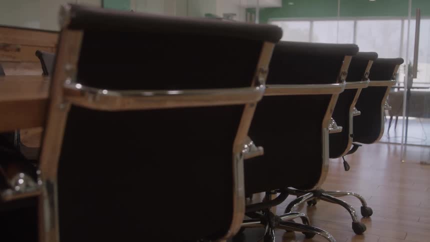 The video is a lowangle tracking shot that moves along the side of a long wooden conference table in a bright office setting a line of matching black executive chairs with metal frames and wheels is visible positioned neatly around the table