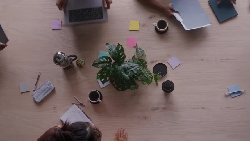 The video is a highangle shot capturing a group of people working together at a large wooden conference tablethe table is cluttered with laptops notebooks clipboards papers coffee cups and a potted plant at the center indicating a brains