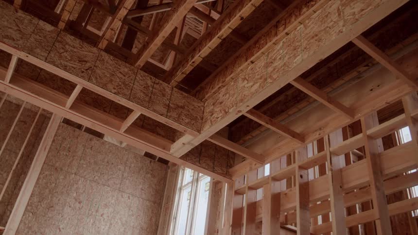 An upwardlooking shot orbits slowly inside an unfinished house revealing the complex wooden framework of studs beams and osb sheathing