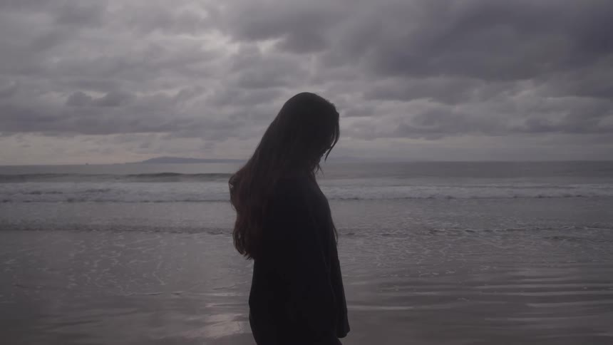 Woman stands on the shore as the waves roll towards her on an overcast autumn day