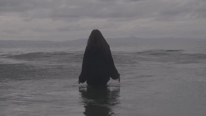 A woman in a dark cloak stands waistdeep in a gray calm sea under heavy clouds facing away with a solitary somber mood