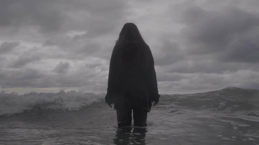 A silhouetted figure of a woman with long hair stands waist deep in the ocean as waves crash around her under a dark cloudy sky