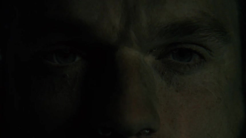 A closeup of a mans face in darkness shows his eyes slowly opening into a stare with his face mostly in shadow