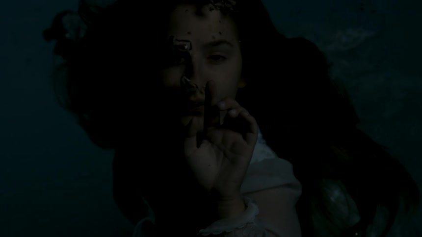 A woman with long dark hair is seen in an extremely dark underwater setting she is wearing a lightcolored garment and her hand is raised toward her face as bubbles float around her