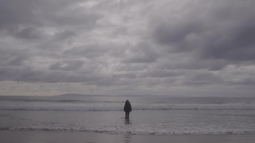 A single figure wearing a dark coat stands alone in the ocean facing the water with their back to the camera the scene is dominated by a vast gloomy sky filled with dark gray clouds giving the environment a dramatic and somber mood