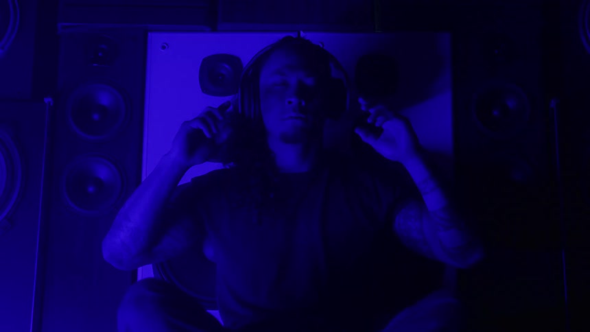 A man with his hair tied back is sitting directly in front of a wall of large stereo speakers illuminated by rapidly flashing red and blue lights he puts on a pair of headphones closes his eyes and appears to be deeply immersed in the musi