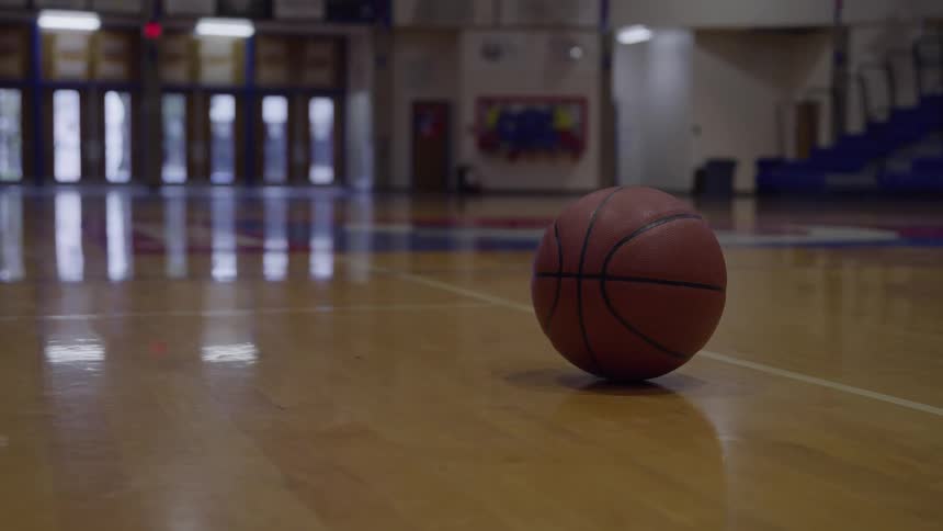 A lone basketball sits silently on a polished wooden court reflecting the soft light of the empty gymnasium and its distant outoffocus bleachers
