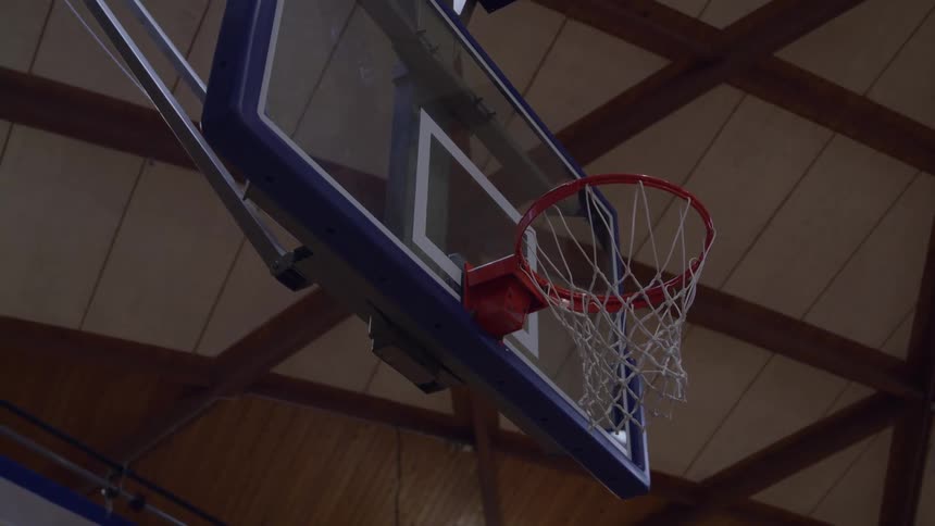 A redrimmed basketball hoop and net hang from a glass backboard inside a gym with a timber ceiling