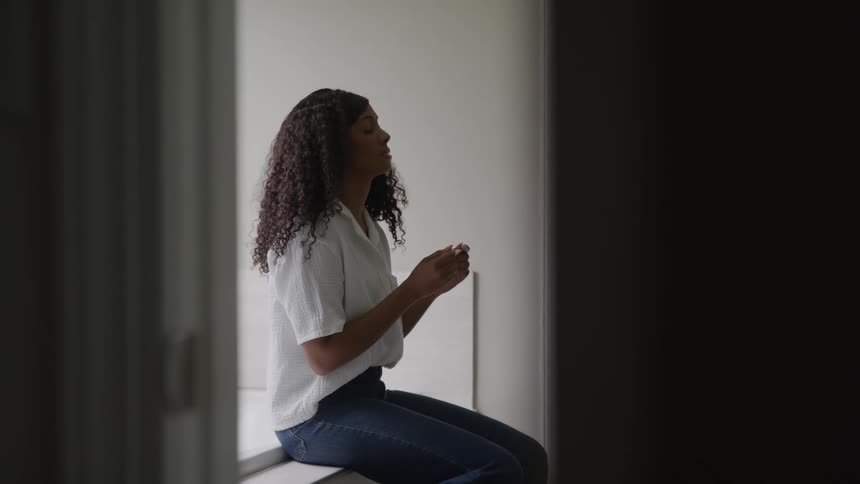 The video is an interior shot of a person with long curly hair sitting sideways on a window ledge looking down at something in their handsthey are bathed in natural light from a large window to their side creating a highcontrast silhouette