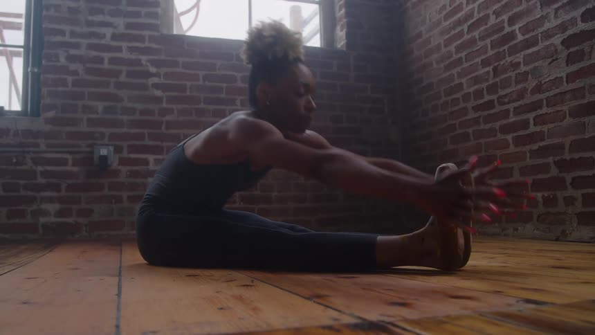 A dancer is performing a floor stretch in a studio with exposed brick walls and large industrial windows she is wearing a dark formfitting unitard while stretching her legs out on the wooden floor