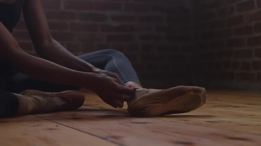 A closeup shot shows a dancer sitting on a wooden floor and tying the ribbonsstraps of a beige dance shoe the background features a dark brick wall in a dimly lit studio space