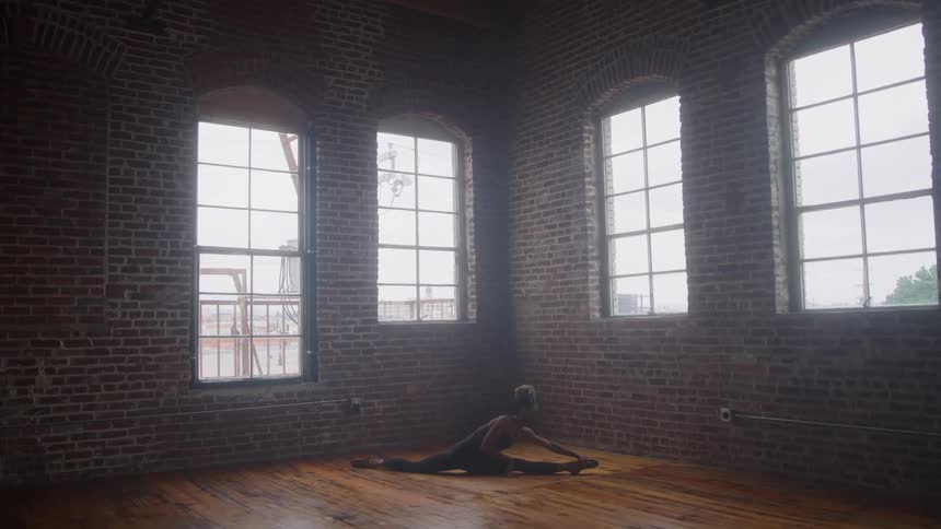 A full shot of a dancer practicing in a loft with a red brick wall and arched windows she is sitting on the wooden floor in a split position before gracefully leaning down and stretching her torso to the floor