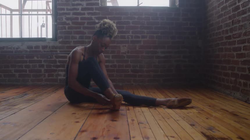 A young female dancer with her hair in a bun is sitting on a wooden floor in a studio with a brick wall background she is wearing dark practice clothes and attentively adjusting her dance shoe