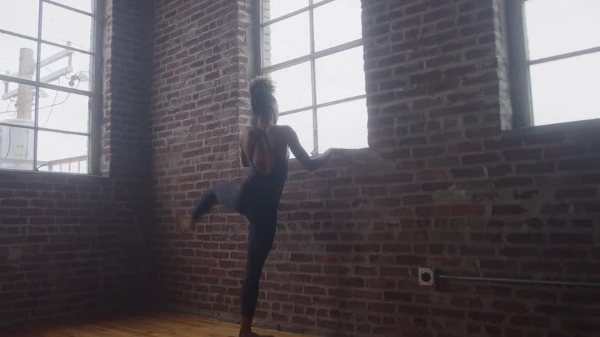 A female dancer is practicing a ballet movement likely arabesque or dgag in a rustic studio with a red brick wall and large windows she is facing the window and lifting one leg high behind her while using the window ledge for balance