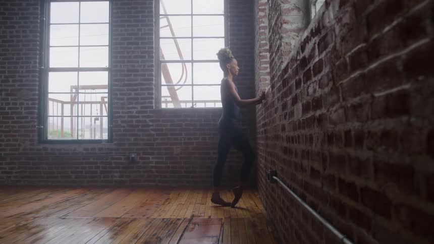 A young black female ballet dancer stretches beside a window at midday inside a room with a wooden floor