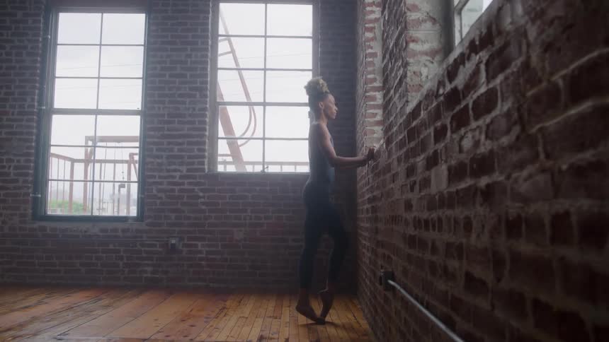 A dancer with hair pulled up into a bun wearing dark leggings and a dark top stands en pointe and presses her hands against a rough brick wall in a rustic loft space with large industrial windows