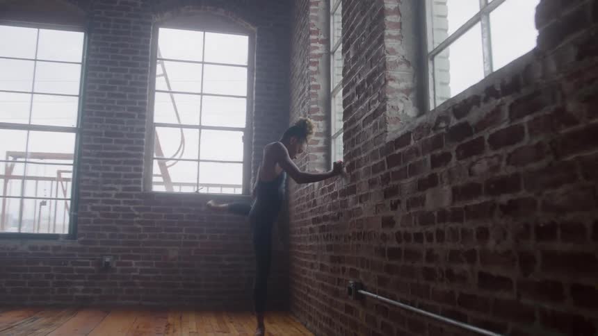A female dancer is seen practicing in a loft studio with a red brick wall and large industrial windows she begins in a vertical split holding her leg high then gracefully lowers her leg and leans forward against the wall