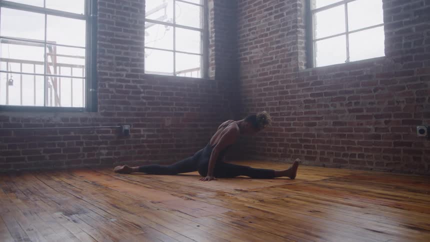 A woman wearing dark athletic wear sits on a wood floor and holds a full split exposed brick walls surround the space and natural light streams in through large industrial windows
