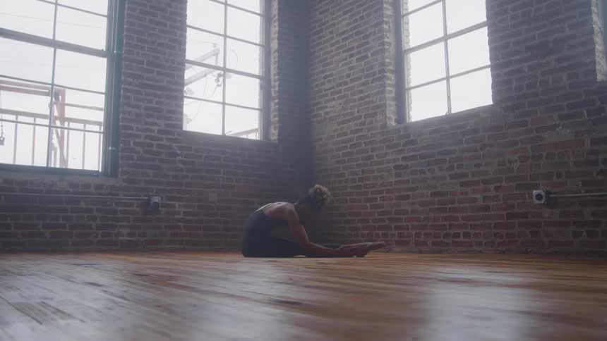 A fullframe shot shows a dancer in an industrialstyle studio with a wooden floor and brick walls she is gracefully executing a deep forward fold stretch from a seated position before slowly beginning to rise and transition to the next move