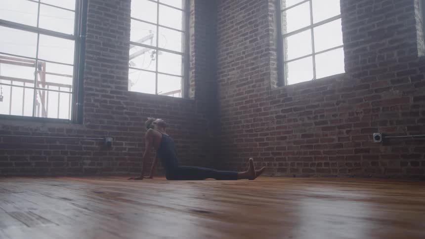 A young female ballet dancer sits on a wooden floor at midday inside an empty room with multiple windows stretching her feet