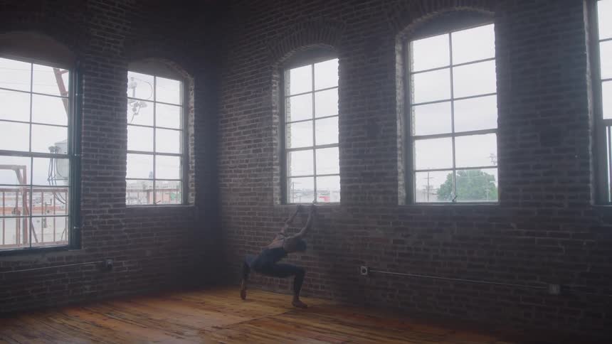 A dark industrial space with brick walls and arched windows features a woman in dark clothing performing a ballet dance move on a wooden floor in a gloomy atmosphere