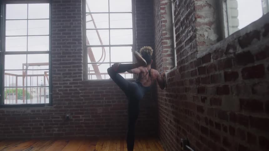 A skilled female dancer is performing a standing split holding her highraised leg with both hands in a studio with exposed red brick walls the shot highlights the dancers flexibility and the contrast with the large industrial windows in th