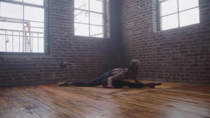 A fit woman in black workout clothes performs a split stretch on a wooden floor inside a bright industrial space with red brick walls and large windows
