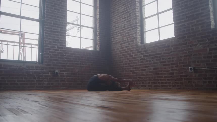 A black female sits on a wood floor in a studio stretching her arms and legs