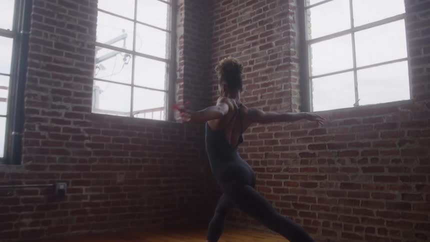 A wide shot captures a dancer in a dark outfit performing an arabesque on a wooden floor in a studio with large arched windows and exposed brick walls the dancer holds a balanced pose with one leg extended as soft diffused light comes in through the win