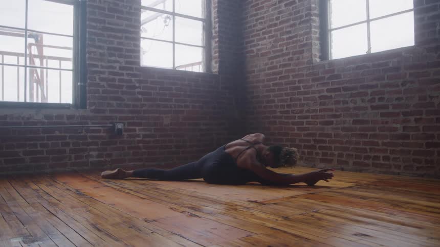 A ballet dancer in black tight clothing poses gracefully on a wooden floor indoors at midday