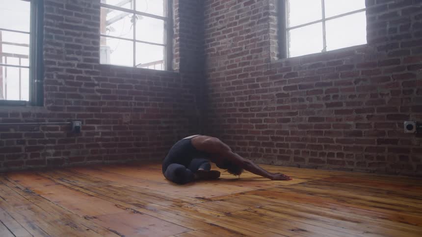 A black female is stretching while sitting in a studio with wood floors