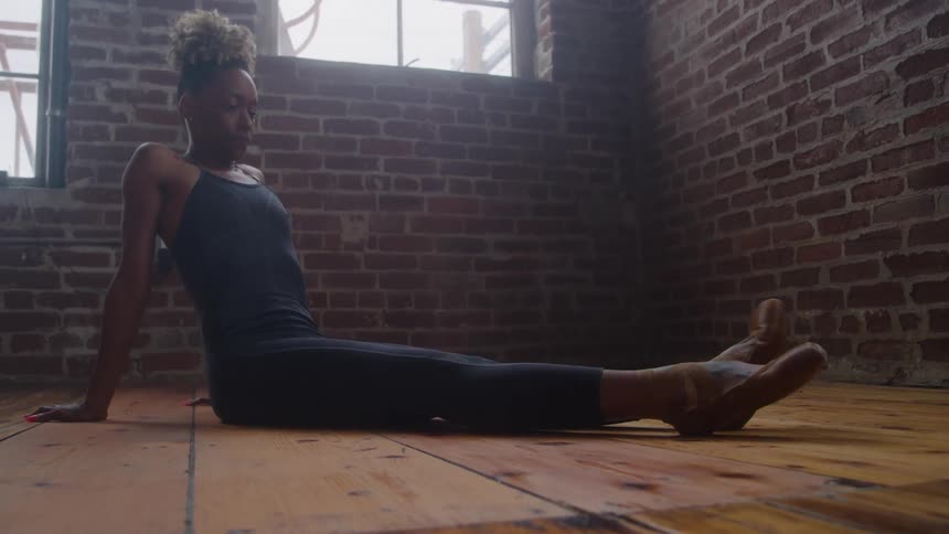 A female dancer is sitting on a wooden floor in a spacious studio leaning back slightly with her legs stretched out in front of her she is dressed in dark dancewear with a red brick wall and tall windows visible in the background