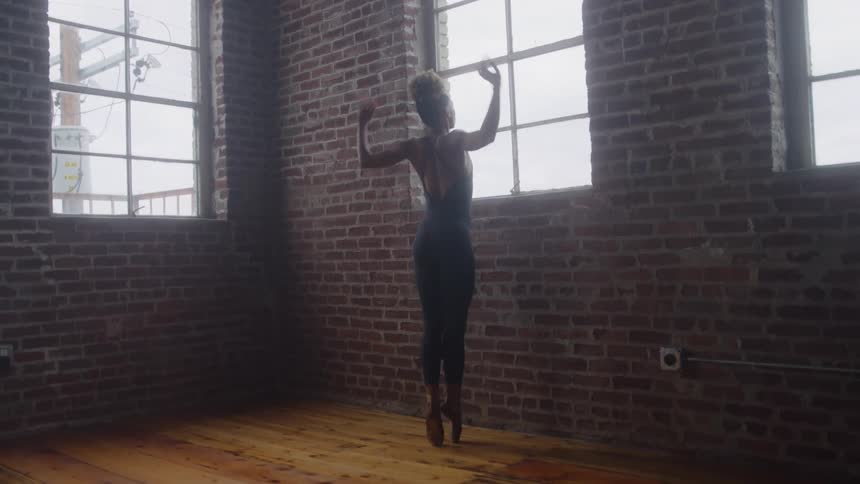 A young woman ballet dancer practices her moves alone at midday in a spacious empty room filled with natural light streaming through multiple tall windows