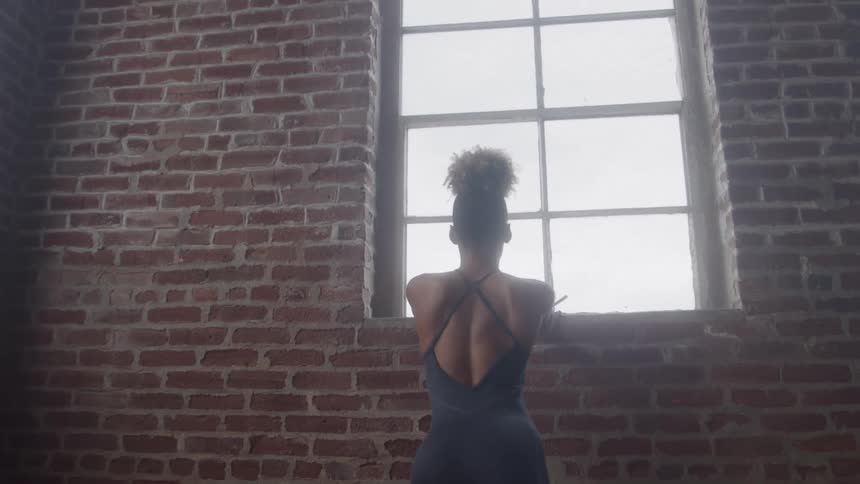 A black female ballet dancer in black tight clothing and ballet shoes stretches beside a wall at midday inside a large room with a wooden floor