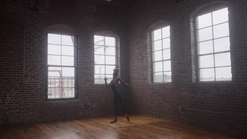 A ballet dancer practices her moves at midday in an empty room with a wooden floor dressed in black tight clothing