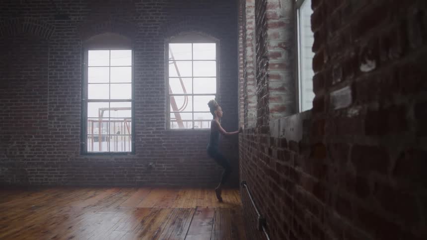 A female ballet dancer stretches beside a brick wall at midday in a large empty room with a wooden floor