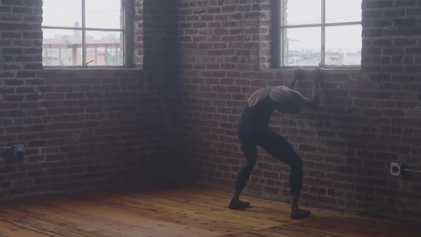 A young female ballet dancer holds the edge of a window and stretches her back bending back and forth at midday inside a room with a wooden floor