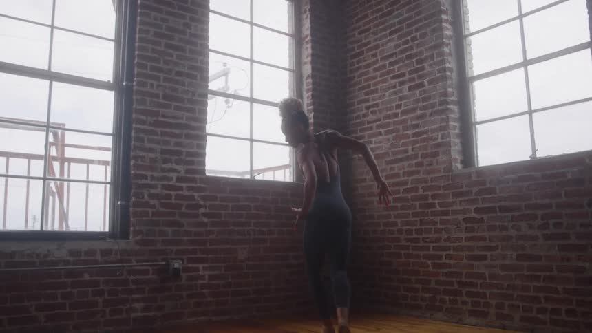 A young woman in tight clothing dances and practices her ballet moves alone in an empty room at midday