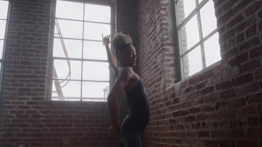 A female ballet dancer practices her dance moves alone beside a window in a quiet room at midday