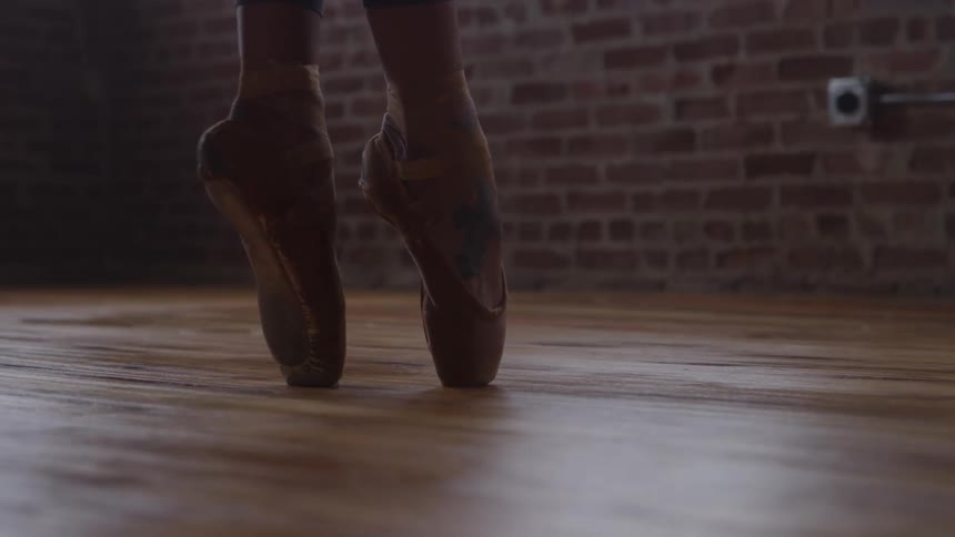 A persons feet in ballet shoes dance on tiptoe across a wooden floor indoors at midday