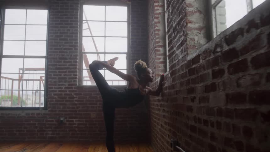 A young ballerina leaning against a wall with one hand and stretching her leg with the other
