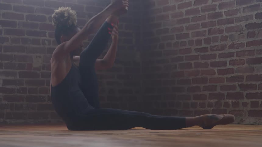 A woman wearing a dark leotard sits on a wood floor with one leg extended she lifts and lowers the other leg in a controlled movement and a red brick wall serves as the background under dramatic lighting
