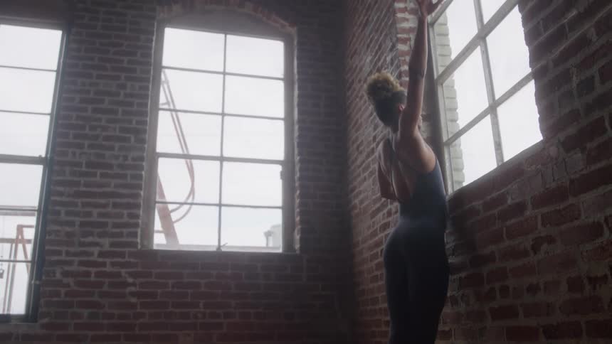 The video shows a dancer in dark activewear performing dynamic and expressive movements near a wall in a spacious room the setting is an industrial or loft space with tall multipane windows and exposed red brick walls