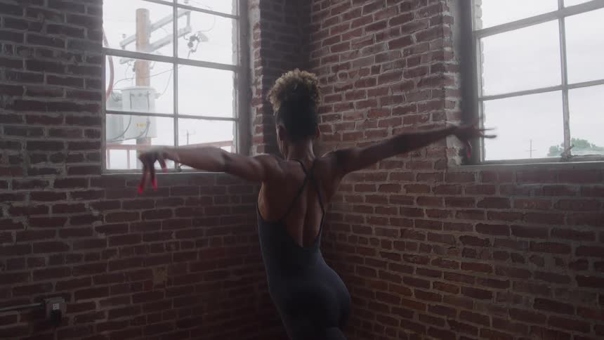 A female ballet dancer practices her moves indoors at midday positioned beside a building wall