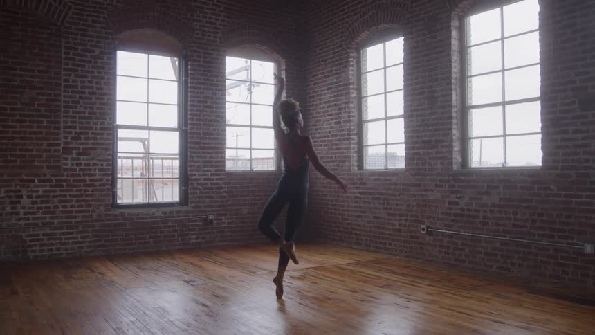 A female dancer practices by herself in an empty room with large windows