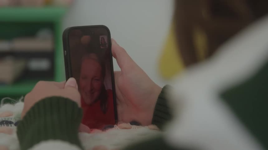 A person holds a phone viewing a video call with a smiling whitehaired elderly man the person holding the phone is wearing a green and white knitted sweater and is sitting indoors