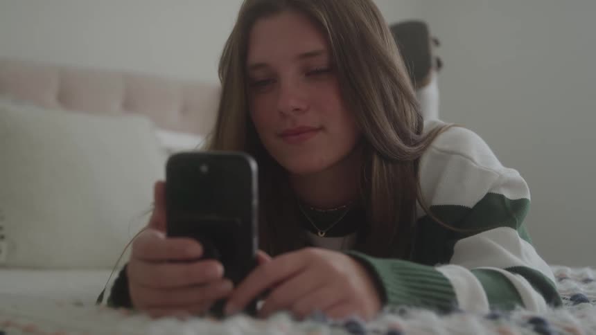 A young teenage girl lies on her bed smiling as she looks at her mobile phone and nods her head with enjoyment