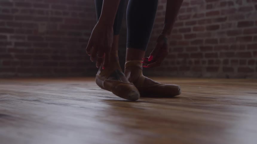 A female wearing ballet shoes stands indoors on a wooden floor at midday poised in a quiet room